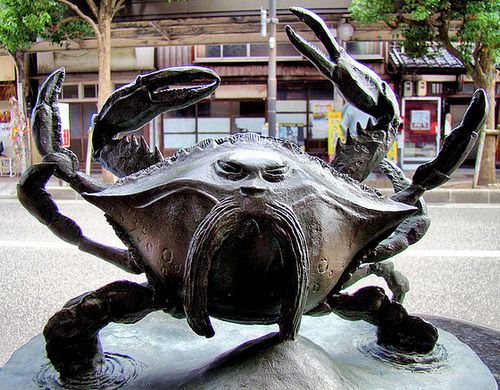 This image shows a large bronze crab statue. The crab is in a rather offensive stance, with its claws raised while standing on its back two claws. The back of the crab facing the camera seems to have a face with two eyes a large mustache.