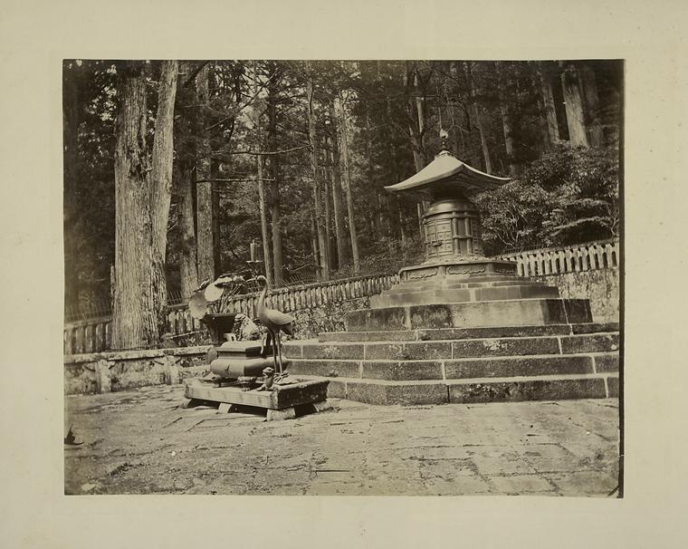 This a sepia tone photo of the tomb of Tokugawa Ieyasu. His tomb rests upon a set of small brick stairs. In front of said stairs appears to be a small statue of what appears to be a heron. In the back we see woods towering over the tomb itself.